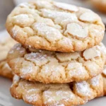 A beautifully arranged plate of golden-brown Italian almond cookies, sprinkled with powdered sugar, set on a rustic wooden table. The background features soft natural light filtering through a kitchen window, with hints of almond flour, whole almonds, and a small bowl of egg whites nearby. The cookies are slightly glossy, showcasing a delicate texture, inviting feel, and warm homemade appeal. -winsome recipes