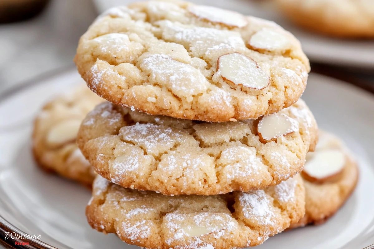 A beautifully arranged plate of golden-brown Italian almond cookies, sprinkled with powdered sugar, set on a rustic wooden table. The background features soft natural light filtering through a kitchen window, with hints of almond flour, whole almonds, and a small bowl of egg whites nearby. The cookies are slightly glossy, showcasing a delicate texture, inviting feel, and warm homemade appeal. -winsome recipes