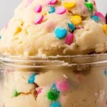 A jar filled with edible sugar cookie dough topped with colorful sprinkles and surrounded by edible sugar cookie dough.