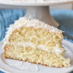 A slice of Ina Garten's coconut cake on a white plate, topped with shredded coconut and a light frosting.